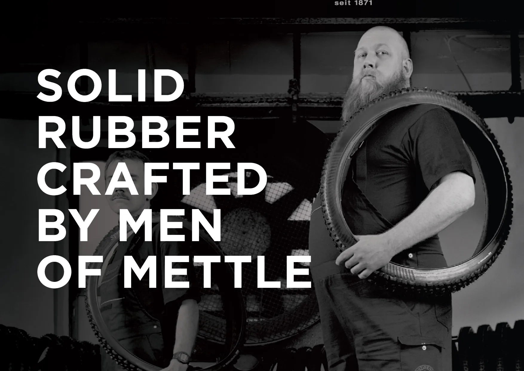 Black-and-white campaign photo of two men in a tyre workshop, one carrying a tyre on his shoulder, with bold white campaign type.