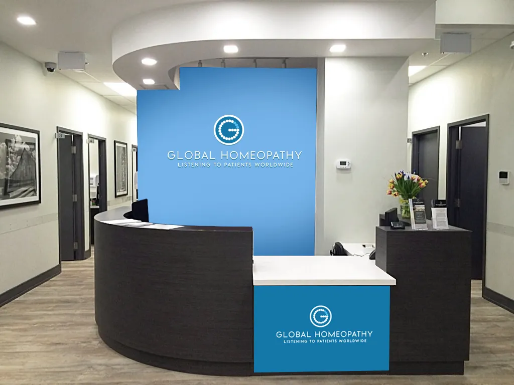 Clinic reception mockup: a curved dark-wood desk with a blue Global Homeopathy branded wall panel behind it.